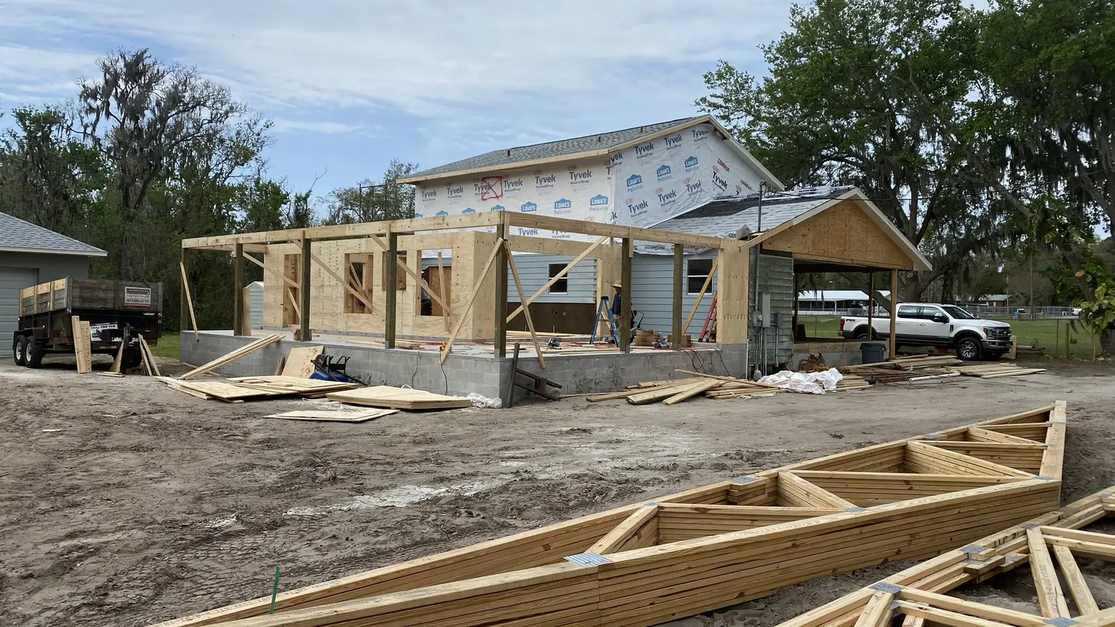 A room addition under construction on a block stem wall alongside an existing house, with roof trusses stacked on the lot ready to set